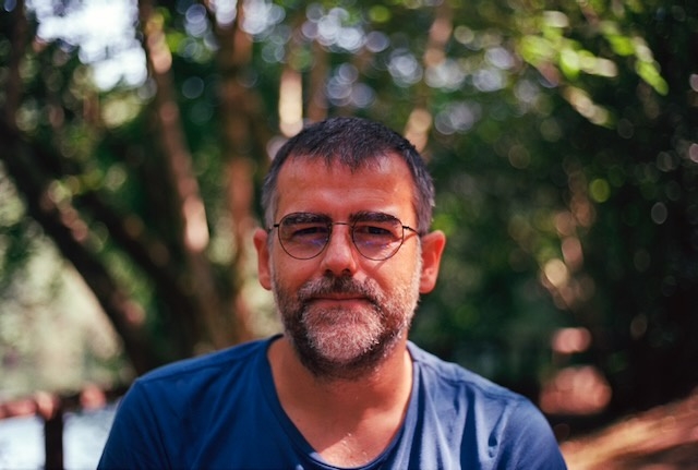 Una fotografía de retrato donde se ve a Ricard Ramon, vestido con una camiseta azul, barba de unos días muy característica, gafas de montura discreta de metal y el pelo corto y moreno. Al fondo, un paisaje de árboles difuminado.