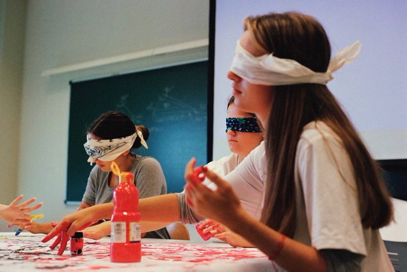 Unas estudiantes, pintan con los dedos sobre un papel en la mesa con los ojos vendados.