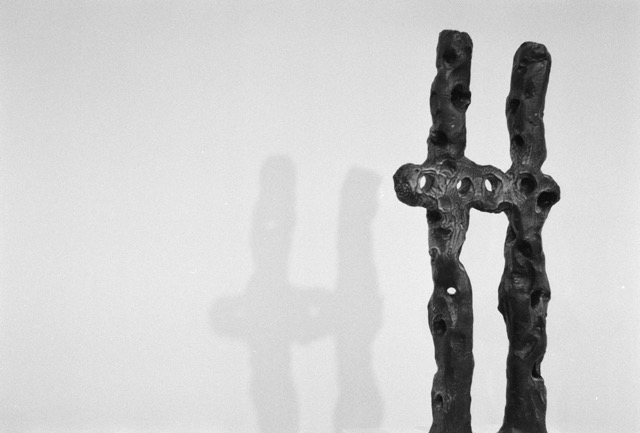 Una escultura de cerámica compuesta por una forma órganica en H, proyectando su sombra contra una pared planca. Foto en vlnaco y negro.