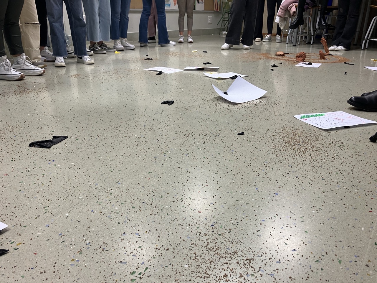 Los pies y las piernas de un grupo de alumnado en círculo en un aula. Sobre el suelo, papeles y otros objetos.