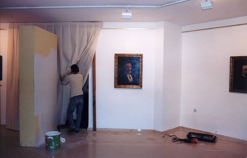 Vista d'una sala d'exposicions amb un quadre penjat i una persona obrint una cortina i treballant en en muntatge.
